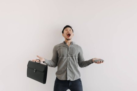 Photo for Asian man with beard wear grey shirt, wow surprised face holding black briefcase and phone, amazed and funny face excited looking at empty space standing isolated over white background wall. - Royalty Free Image