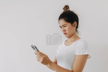 Téléchargez les photos : Femme thaïlandaise asiatique utilisant un téléphone mobile et regardant avec un drôle de visage de doute, tenant smartphone par pose suspecte silencieuse, debout isolé sur un mur de fond blanc. Photo de haute qualité - en image libre de droit