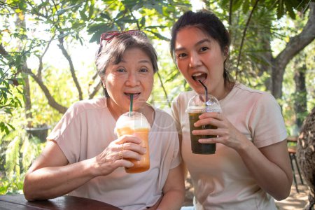 Photo for Happy asian Thai Chinese elder mother and daughter drinking cold beverages together and sitting at cafe in garden, spending good time looking at camera. - Royalty Free Image