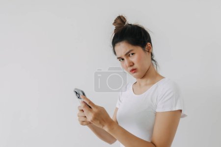 Téléchargez les photos : Femme thaïlandaise asiatique utilisant un téléphone mobile avec un drôle de visage de doute, tenant smartphone par pose suspecte silencieuse, debout isolé sur un mur de fond blanc. - en image libre de droit