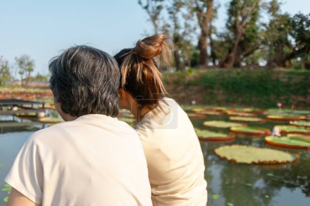 Foto de Vista trasera trasera de la madre anciana tailandesa asiática y su hija sentadas en la orilla junto a la laguna local del lago de flores de loto, ambas mujeres felices sonriendo, viajan de vacaciones. - Imagen libre de derechos