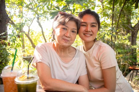 Téléchargez les photos : Asiatique thaïlandais chinois aîné mère et fille câlins et prendre des photos regarder appareil photo, boire des boissons au café jardin, passer du temps ensemble. - en image libre de droit