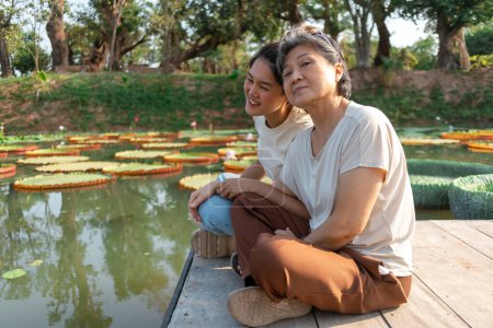 Téléchargez les photos : Vue latérale de la mère et de la fille aînée chinoise asiatique thaïlandaise assise sur la rive par le lac de fleur de lotus lagune locale, les deux femmes souriantes heureuses, voyagent en vacances sous le soleil. - en image libre de droit
