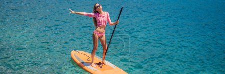 BANNER, LANG FORMAT Junge Frauen haben Spaß beim Stand Up Paddling im blauen Meer in Montenegro. SUP. Mädchen trainiert auf Paddelbrett in der Nähe der Felsen.