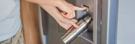 Male hand is pouring cold water and ice cubes in a metal bottle from dispenser of home fridge. BANNER, LONG FORMAT
