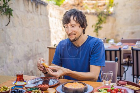 Téléchargez les photos : Un homme qui mange un petit déjeuner turc. Table de petit déjeuner turque. Des pâtisseries. Des légumes. Des olives. Fromages, œufs frits. Confitures, thé en pot de cuivre et verres à tulipes. Grande composition. - en image libre de droit