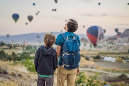 Foto de Turistas padre e hijo mirando globos aerostáticos en Capadocia, Turquía. Feliz viaje en Turquía concepto. padre e hijo en una cima de montaña disfrutando de una vista maravillosa. - Imagen libre de derechos