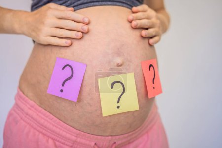 Téléchargez les photos : Choisir le nom du bébé. Femme enceinte confondue avec des points d'interrogation sur des autocollants en papier sur le ventre
. - en image libre de droit