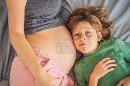Foto de Joven mujer embarazada hermosa y el hijo mayor. El chico lindo apoyó su oreja contra el vientre de su madre. Esperando un bebé en el concepto de familia. Preparar a un niño mayor para uno más pequeño. - Imagen libre de derechos