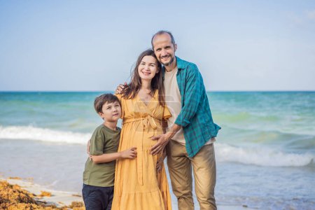Eine liebevolle Familie genießt den tropischen Strand - eine strahlend schwangere Frau nach 40, umarmt von ihrem Mann und begleitet von ihrem erwachsenen Sohn im Teenageralter, die gemeinsam kostbare Momente inmitten der Natur genießt.