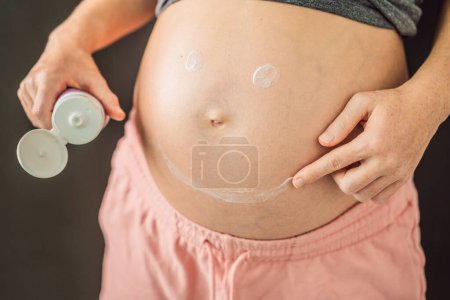 Photo for Positive young pregnant woman in comfortable homewear applying belly butter on her big tummy, woman belly. - Royalty Free Image