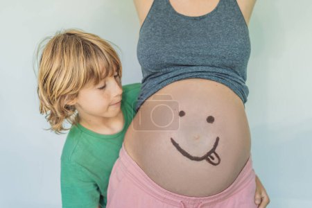 Liebenswerter Moment als Sohn verleiht der Schwangerschaft seiner Mutter einen Hauch von Freude, indem er spielerisch ein lustiges Gesicht auf ihren Babybauch zeichnet und gehegte Erinnerungen weckt.