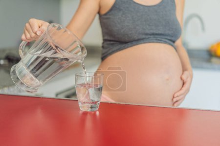 Eine schwangere Frau umarmt die lebenswichtigen Vorteile von Wasser während der Schwangerschaft und steht mit einem Glas in der Küche, um die entscheidende Rolle der Hydratation für das Wohlbefinden der Mutter hervorzuheben..