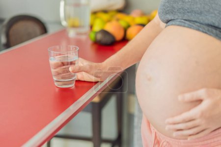 Eine schwangere Frau umarmt die lebenswichtigen Vorteile von Wasser während der Schwangerschaft und steht mit einem Glas in der Küche, um die entscheidende Rolle der Hydratation für das Wohlbefinden der Mutter hervorzuheben..