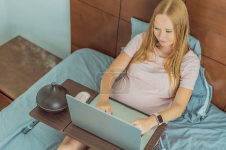 Foto de Mujer embarazada multitarea mejora su espacio de trabajo en casa, utilizando un difusor de aroma para una atmósfera relajante durante el trabajo durante el embarazo. - Imagen libre de derechos