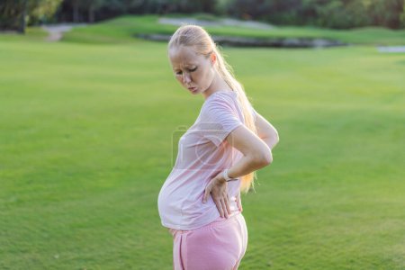 Werdende Frau erlebt Rückenschmerzen im Freien und sucht Linderung und Trost während der Schwangerschaft mit einer sanften Dehnung oder Erholung im Freien.