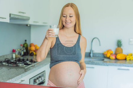 Téléchargez les photos : Pesant le pour et le contre du lait pendant la grossesse, une femme enceinte réfléchie se tient dans la cuisine avec un verre, envisageant la décision d'inclure ou d'éviter le lait pour elle et ses bébés bien - en image libre de droit