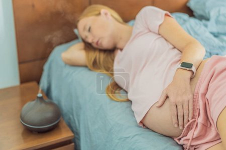 Foto de Mujer embarazada encuentra la relajación en la cama, abrazando la tranquilidad con la ayuda de un difusor de aroma durante un momento de descanso. - Imagen libre de derechos