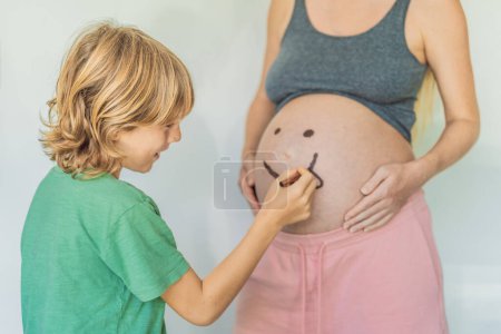 Liebenswerter Moment als Sohn verleiht der Schwangerschaft seiner Mutter einen Hauch von Freude, indem er spielerisch ein lustiges Gesicht auf ihren Babybauch zeichnet und gehegte Erinnerungen weckt.