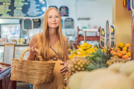 En una tienda de comestibles, una mujer embarazada está junto a un puesto de frutas, rodeada de varios alimentos naturales. Ella está en un espacio público donde el mercado local ofrece alimentos enteros para el comercio