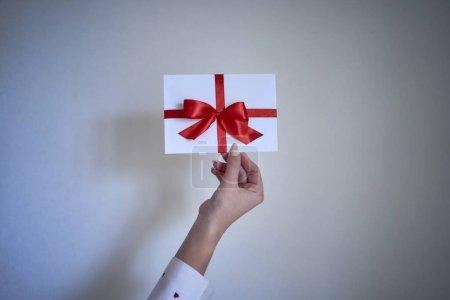 Téléchargez les photos : Un certificat cadeau en couleurs rouge et blanc dans les mains féminines sur un fond de mur blanc - en image libre de droit