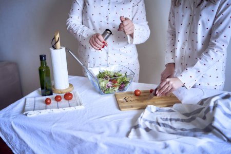 teenage girl and her mother in pajamas are cooking and eating a fresh green and tomato salad together                    
