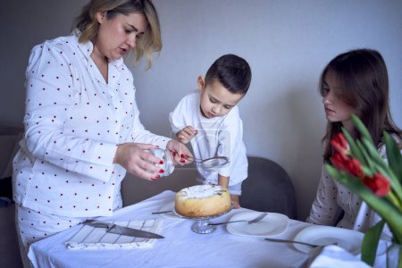         a  family of three, mother, teenage daughter and little son, eating cake in pajamas at a table with tulips                     