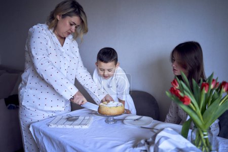 a family of three, mother, teenage daughter and little son, eating cake in pajamas at a table with tulips
