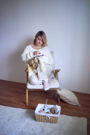 a midsize woman in light clothes embroiders a picture in gold and beige tones while sitting on a white chair in a light room