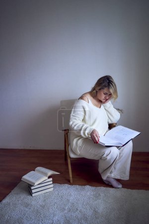   medium-sized woman in light clothes reads a book while sitting in a white chair in a light room                     