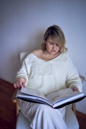   medium-sized woman in light clothes reads a book while sitting in a white chair in a light room                     