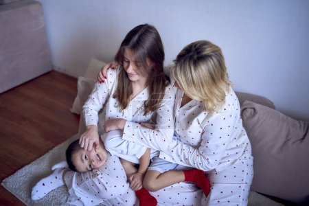 mother and teenage girl in pajamas tickle younger brother on the floor