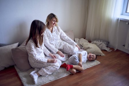 mother and teenage girl in pajamas tickle younger brother on the floor
