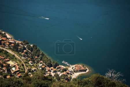             Stadt und die sandige Halbinsel am Ufer des Comer Sees von oben, zwei Boote schneiden durch das türkisfarbene Wasser und hinterlassen eine weiße Spur                    