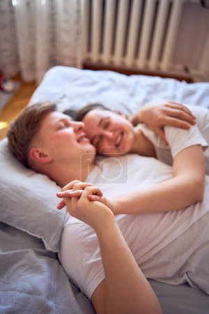 Téléchargez les photos : Un jeune couple sur le lit tenant la main, une fille sur l'épaule d'un garçon, câlins tendres et touches - en image libre de droit