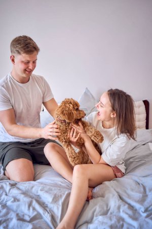 Téléchargez les photos : Couple joue avec une fille cockapoo sur un lit, minimalisme - en image libre de droit