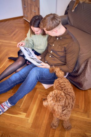 a young couple looking at their wedding photo album by the bed in living room, cockapoo running around                           