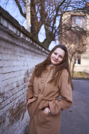                     a girl in a brown coat is happy  near a white wall on a spring day           