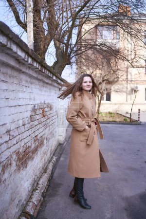                     ein Mädchen im braunen Mantel freut sich an einem Frühlingstag an einer weißen Wand           