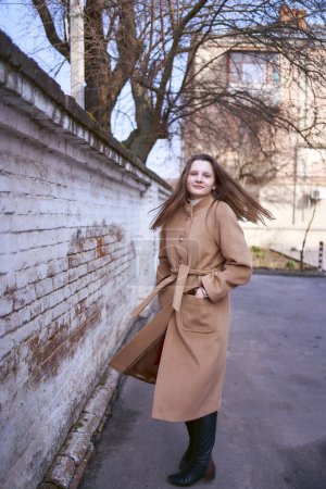                     a girl in a brown coat is happy  near a white wall on a spring day           