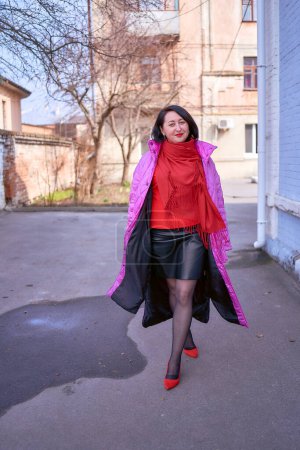 stylish middle age woman in a long bright pink, magenta coat and red shoes walks the streets of the city        
