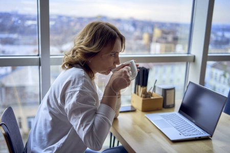         una joven rubia bebiendo café y trabajando en un portátil en un café con ventanas panorámicas y una vista de la ciudad desde arriba                       