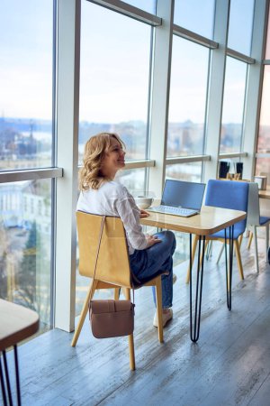  femme en pull et jeans boit du café et travaille sur un ordinateur portable dans un espace de coworking avec des fenêtres panoramiques et une vue sur la ville d'en haut       