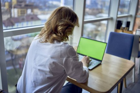 Téléchargez les photos : Femme d'âge moyen travaillant sur un ordinateur portable dans un espace de coworking avec des fenêtres panoramiques et une vue sur la ville d'en haut, ordinateur portable avec un écran vert, clé Chroma - en image libre de droit