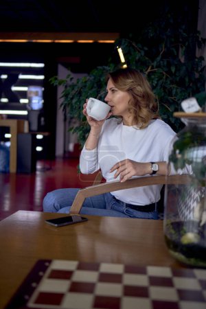   a beautiful middle age woman drinking coffee in a beautiful cafe with greenery and light panels in the background                            