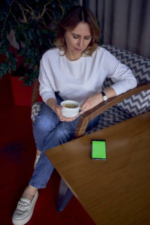 eine schöne Frau mittleren Alters trinkt Kaffee in einem schönen Café mit viel Grün und hellen Tafeln im Hintergrund, ein Telefon mit grünem Bildschirm auf dem Tisch                          