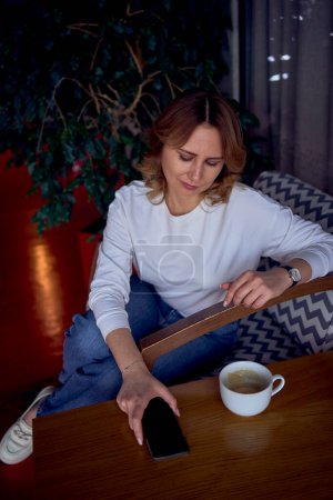 Téléchargez les photos : Une jolie femme d'âge moyen recherche sur son téléphone dans un beau café avec de la verdure et des panneaux lumineux en arrière-plan - en image libre de droit