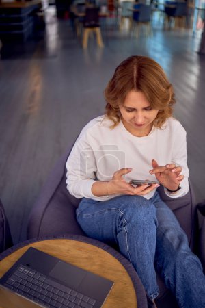  mujer en ropa casual se sienta en una bolsa de frijoles y busca usando el teléfono                   