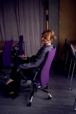        schöne Geschäftsfrau mittleren Alters mit grauem Hemd, breiter Beinhose und schwarzen Stilettos sitzt im lila Bürostuhl in einem modernen Arbeitsbereich                        