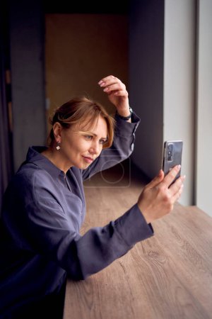                beautiful middle age business woman wearing gray shirt, wide leg pants and black stilettos fix her hair looking into phone camera in modern workspace                                               